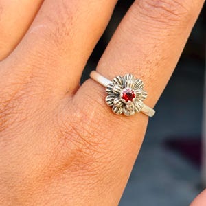 May include: A silver ring featuring a flower design with a red gemstone center. The ring is worn on a finger, showcasing the intricate details of the floral pattern and the vibrant red stone.