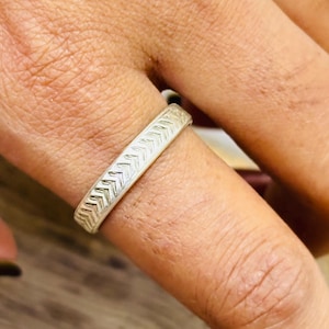 Puede incluir: Un anillo plateado con un patrón de chevrón se muestra en un dedo. El anillo tiene una superficie texturizada con una serie de pequeñas formas repetidas en forma de flecha. El anillo es un diseño de banda simple.