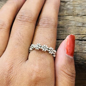 Puede incluir: Un anillo de tono plateado que presenta una banda continua de pequeñas formas florales en relieve similares a margaritas. El metal tiene un acabado ligeramente oxidado en las áreas hundidas entre los pétalos, resaltando el patrón floral.
