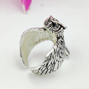 Könnte beinhalten: Ein silberfarbener Metallring in Form einer Eule mit ausgebreiteten Flügeln. Die Eule weist detaillierte Federstrukturen und einen markanten Kopf mit definierten Augen auf. Die Ringschiene wird durch die Flügel der Eule gebildet, was ein offenes Design ergibt.