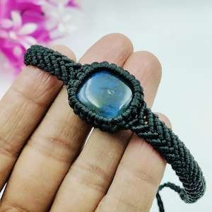 Peut inclure: Bracelet en macramé noir orné d'une pierre précieuse bleue carrée et irisée. La pierre est sertie dans un cordon noir tissé, avec des détails de nœuds complexes. Le bracelet présente un motif tressé sur les côtés.