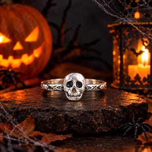 Op de afbeelding: Een zilverkleurige ring met een centraal schedelmotief met donkere, verdiepte details. De ringband is voorzien van een herhalend geometrisch patroon aan de zijkanten, wat een gestructureerd uiterlijk geeft aan het metalen oppervlak.