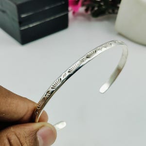 Puede incluir: Una pulsera brazalete fina de metal color plata que presenta una serie de pequeños patrones estampados en forma de abanico a lo largo de la superficie exterior. La pulsera tiene un diseño abierto con extremos lisos y redondeados.