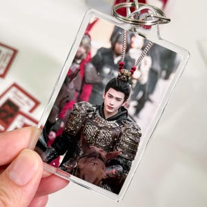 May include: A rectangular clear acrylic keychain featuring a printed image of a person with dark hair styled in a high bun, wearing ornate dark metal armor with intricate detailing, and sitting atop a horse with a small mark on its forehead.