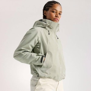 Könnte beinhalten: Hellgrüne Kapuzenjacke mit Reißverschluss und Taschen. Die Jacke wird mit cremefarbenen Hosen getragen. Das Model trägt die Jacke und schaut in die Kamera. Die Jacke hat einen hohen Kragen und eine Kapuze.