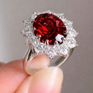May include: A silver ring with a large, oval, deep red gemstone, encircled by smaller, round, clear gemstones. The ring's band is polished silver, and the setting is detailed. The ring is a piece of jewellery.