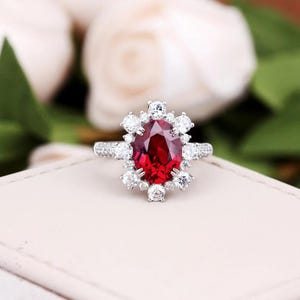 May include: A silver-coloured ring featuring a large, oval-cut deep red gemstone at the centre. The central stone is surrounded by a halo of twelve smaller, round-cut clear white stones, with additional small clear stones set along the ring band.