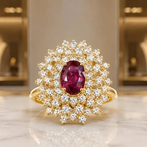 Peut inclure: Une bague en or avec un rubis rouge foncé taille ovale au centre, entouré de deux halos concentriques de petits diamants blancs ronds et clairs. La monture est en or jaune poli, formant un motif en grappe inspiré d'une fleur.