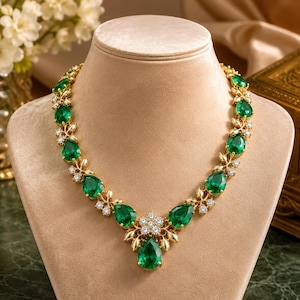 Puede incluir: Un collar de tono dorado con una serie de piedras verde esmeralda en forma de pera que se alternan con grupos de pequeñas piedras blancas redondas y transparentes. El punto central es una piedra verde más grande rodeada de piedras blancas.