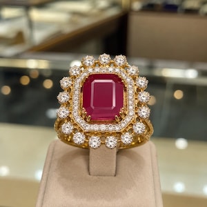 Puede incluir: Un anillo de oro con un gran rubí rectangular de color rojo intenso en el centro, rodeado por un doble halo de pequeños diamantes blancos redondos y claros. El engaste es de oro amarillo pulido con un diseño de borde festoneado.