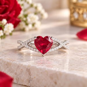 Puede incluir: Un anillo de tono plateado con una prominente piedra preciosa roja en forma de corazón en el centro. La banda presenta un diseño entrecruzado y está incrustada con pequeñas piedras claras y brillantes a lo largo de la montura metálica.