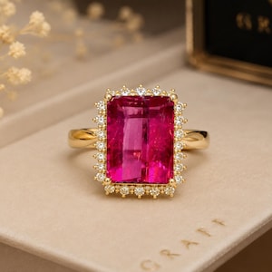 Puede incluir: Un anillo de oro con una gran piedra preciosa rectangular de color rosa vibrante en el centro. La piedra central está rodeada por un halo de pequeños diamantes blancos redondos engastados en oro. El aro del anillo es de oro amarillo pulido.