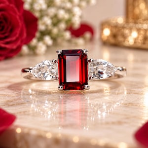 May include: A silver-toned ring featuring a large, rectangular, emerald-cut deep red gemstone at the centre, flanked by two clear, pear-shaped stones on either side, all set on a polished metal band with a four-prong setting for the centre stone.