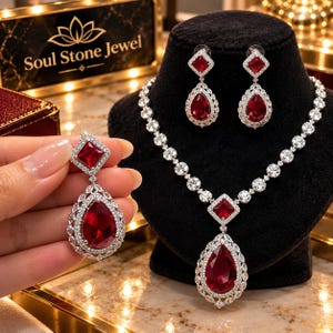 Puede incluir: Un conjunto de joyería que incluye un collar y pendientes a juego con piedras de color rojo intenso en forma de pera y corte cuadrado rodeadas de cristales pavé transparentes. El collar tiene un colgante central. Texto Soul Stone Jewel.