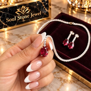 May include: A jewellery set featuring a tennis necklace and matching drop earrings, each set with oval-cut deep red stones and clear round accent stones. The necklace and earrings are displayed on a dark red velvet tray near a Soul Stone Jewel sign.