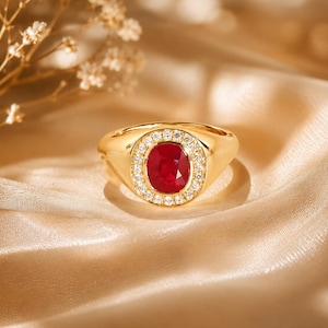 Puede incluir: Un anillo de sello de oro con una piedra preciosa de rubí rojo intenso de corte ovalado en el centro. El rubí está rodeado por un halo de pequeños diamantes blancos de corte redondo engastados en la banda de oro. El anillo tiene un acabado pulido.