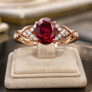 Puede incluir: Un anillo de oro rosa con una gran piedra preciosa de color rojo intenso de talla ovalada en el centro, flanqueada por pequeños diamantes blancos redondos engastados en la banda. La banda de metal presenta un diseño intrincado y entrelazado.