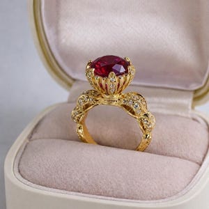 Puede incluir: Un anillo de oro con una gran gema roja facetada. El anillo tiene un diseño detallado y ornamentado con pequeñas piedras claras de acento. El anillo se muestra en un joyero crema con interior rosa.