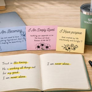 May include: An open notebook with handwritten text and three colorful cards with inspirational quotes. A green Stanley tumbler, a lit candle, a pen, and a laptop are also on the wooden surface.