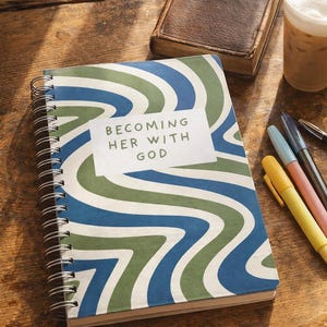 Puede incluir: Un cuaderno con espiral con un diseño ondulado azul, verde y blanco y el texto "BECOMING HER WITH GOD". Una Biblia encuadernada en cuero, bolígrafos y una bebida están sobre una mesa de madera.