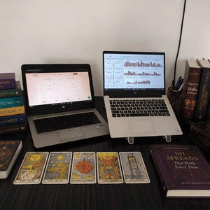 Professional astrology and tarot reading workspace featuring two laptops on a dark wooden desk surrounded by tarot cards, tarot decks, and astrology books. The setup includes Vedic astrology reference books, tarot guidebooks, mystical decor, and displayed tarot cards including The Sun, The Star, Wheel of Fortune, The Lovers, and Ace of Cups, creating a spiritual and modern divination environment.