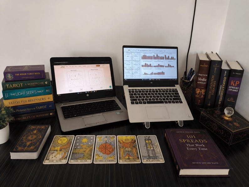 Professional astrology and tarot reading workspace featuring two laptops on a dark wooden desk surrounded by tarot cards, tarot decks, and astrology books. The setup includes Vedic astrology reference books, tarot guidebooks, mystical decor, and displayed tarot cards including The Sun, The Star, Wheel of Fortune, The Lovers, and Ace of Cups, creating a spiritual and modern divination environment.