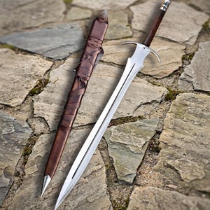 May include: A silver-toned metal sword with a pointed blade, a crossguard, and a dark brown wooden handle with a metallic pommel. Beside the sword lies a matching dark brown leather scabbard featuring intricate leather strap detailing.