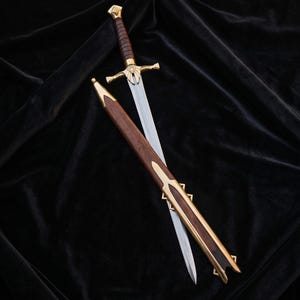 Può includere: Una spada in acciaio con impugnatura rivestita in pelle marrone e una guardia dorata con un motivo ad aquila. La spada è accompagnata da un fodero in legno abbinato con finiture in metallo dorato sulla punta e sull'imboccatura, posizionata in diagonale.