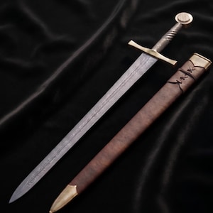 May include: A steel sword with an etched blade, a gold-toned crossguard, and a textured handle with a round pommel. Beside the sword lies a brown leather scabbard featuring a gold-toned metal tip and decorative cross-lacing near the opening.