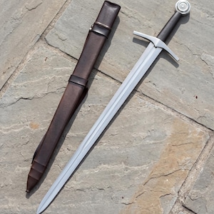 Puede incluir: Una espada de acero con hoja de doble filo, guarda recta y empuñadura negra rematada con un pomo circular de plata que presenta un emblema central. Junto a la espada hay una vaina de cuero marrón oscuro con correas de cuero a juego.