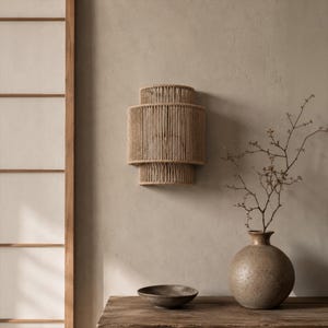 May include: A wall-mounted light fixture featuring a tiered, cylindrical shade constructed from tightly wound natural beige rope. The fixture has a distinct three-section design with a wider central band and narrower top and bottom sections.