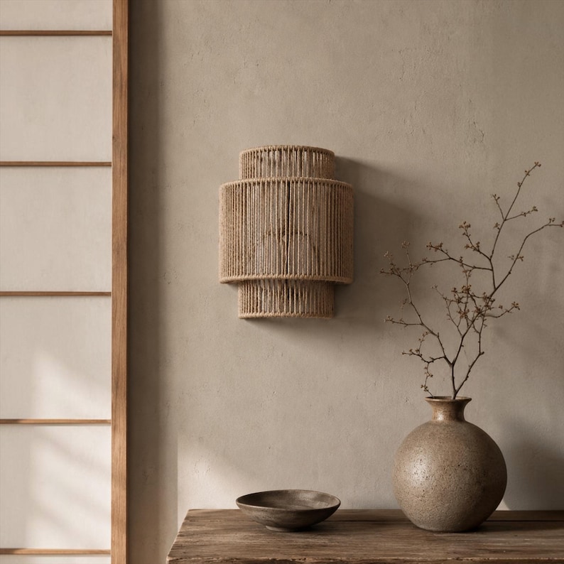 May include: A wall-mounted light fixture featuring a tiered, cylindrical shade constructed from tightly wound natural beige rope. The fixture has a distinct three-section design with a wider central band and narrower top and bottom sections.