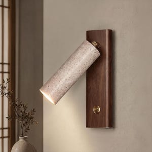 May include: A wall-mounted light fixture featuring a rectangular dark wood backplate and a cylindrical, textured beige stone lamp head. The lamp head is attached to the backplate with a brass-colored hinge and includes a small brass power button.