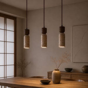 Puede incluir: Tres lámparas colgantes cilíndricas que cuelgan de finos cables oscuros. Cada luminaria presenta una sección superior de madera marrón oscuro lisa conectada a una base de piedra beige texturizada con una superficie porosa y luz cálida.