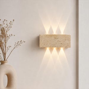 Op de afbeelding: Een rechthoekige wandlamp gemaakt van beige poreuze steen met een getextureerd oppervlak. Het armatuur straalt licht uit in drie duidelijke stralen naar boven en drie stralen naar beneden, wat een symmetrisch verlichtingspatroon op de muur creëert.