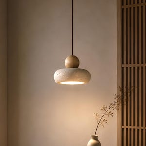 Peut inclure: Un luminaire suspendu moderne avec un abat-jour texturé de couleur claire et une sphère en bois au-dessus. La lumière est suspendue à un cordon foncé. Un vase et un bol sont posés sur une table en bois.