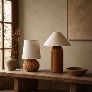 Peut inclure: Deux lampes de table avec des bases en rotin tressé marron et des abat-jours coniques en tissu blanc cassé. La lampe de gauche a une base arrondie et bulbeuse, tandis que celle de droite a une base cylindrique plus haute sur une surface en bois.
