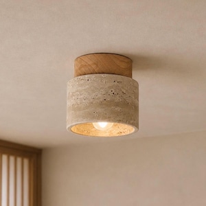 May include: A modern ceiling light fixture. It features a cylindrical shade made of natural stone, topped with a wooden element. The light emits a warm glow. The fixture is mounted on a neutral-toned ceiling. The design is minimalist.