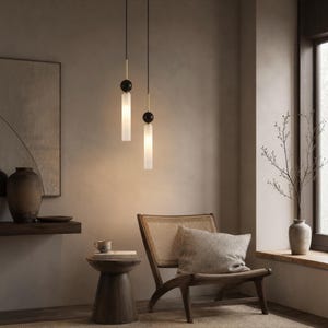 Puede incluir: Dos lámparas colgantes cilíndricas suspendidas de finos cables negros. Cada luminaria cuenta con un conector esférico negro mate, una varilla de metal dorado y un cilindro de vidrio blanco esmerilado que emite una luz cálida y suave.