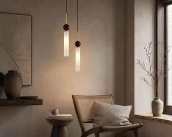 May include: Two cylindrical pendant lights suspended from thin black cords. Each fixture features a matte black sphere connector, a gold-toned metal rod, and a frosted white glass cylinder that emits a soft, warm glow downward into the room.