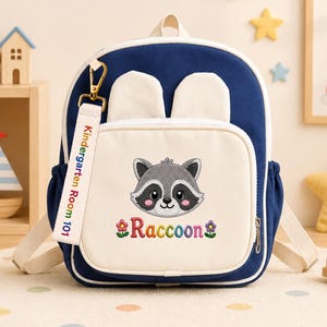 K&ouml;nnte beinhalten: Ein marineblauer und cremefarbener Rucksack mit einem zentralen gestickten Waschb&auml;rengesicht, dem Text "Raccoon" mit Blumensymbolen und einem wei&szlig;en Stoffanh&auml;nger mit der Aufschrift "Kindergarten Room 101". Der Rucksack hat zwei kleine Ohren.
