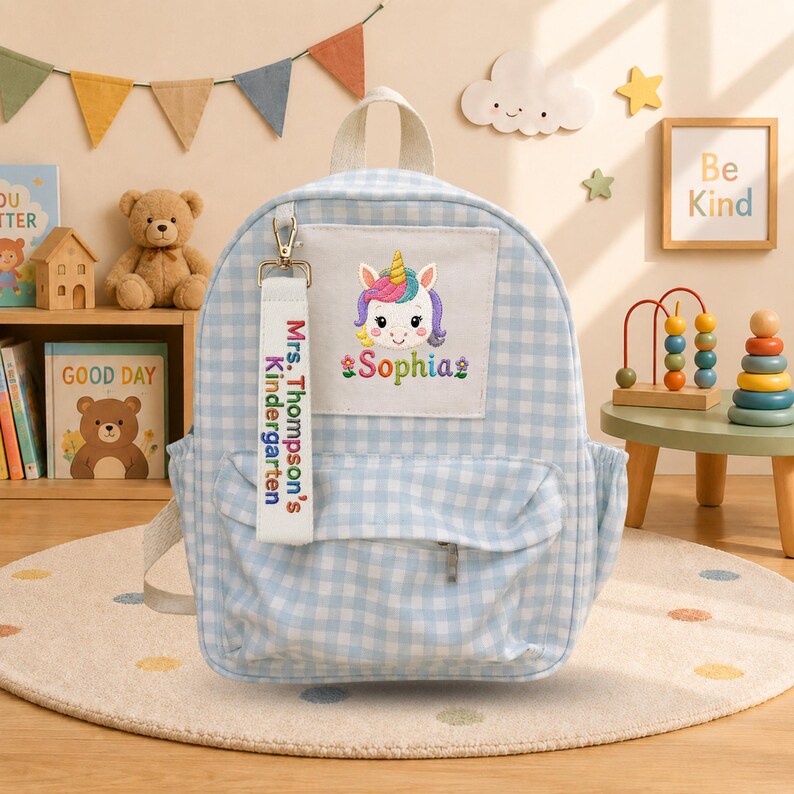 K&ouml;nnte beinhalten: Ein hellblau-wei&szlig;er Rucksack mit Vichy-Karo-Muster und einer wei&szlig;en Vordertasche, die ein gesticktes Einhorn und den Namen Sophia zeigt. Ein wei&szlig;er Stoffanh&auml;nger ist befestigt, der mit dem Text Mrs. Thompson's Kindergarten in bunten Buchstaben bedruckt ist.