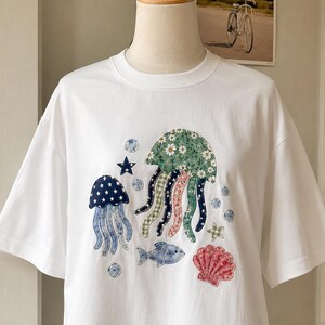 May include: A white short-sleeved t-shirt featuring a colorful fabric appliqué design of two jellyfish, a fish, and a seashell. The jellyfish are decorated with floral, star, and gingham patterns in shades of green, blue, and pink.