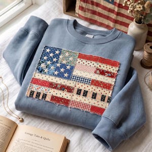 May include: A dusty blue crewneck sweatshirt featuring a rectangular patch on the chest. The patch is a patchwork design of the American flag, constructed from various red, white, and blue fabric scraps with floral, gingham, and star patterns.