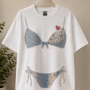 May include: A white short-sleeve t-shirt featuring a graphic print of a two-piece bikini set. The bikini top and bottom are composed of patchwork patterns in blue gingham, floral, and solid light blue fabric, with a small red heart accent.