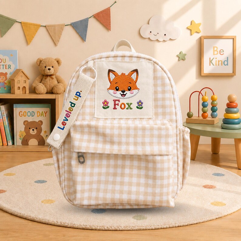 K&ouml;nnte beinhalten: Ein beigewei&szlig;er Rucksack mit Vichy-Karo-Muster, bestickt mit einem Fuchskopf, dem Schriftzug "Fox" und zwei kleinen Blumen. Ein seitlicher Riemen tr&auml;gt den Text "Leveled up." Der Rucksack verf&uuml;gt &uuml;ber eine Vordertasche und einen Tragegriff.