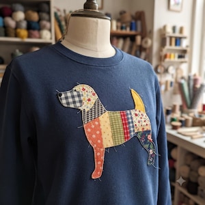 May include: A navy blue crewneck sweatshirt featuring a large, central applique of a dog. The dog is constructed from various fabric patches in different patterns, including gingham, floral, and striped prints in shades of red, green, and yellow.
