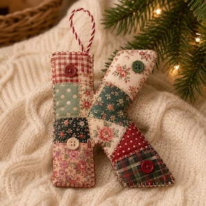 May include: A fabric letter K ornament featuring a patchwork design of various floral, polka dot, and plaid patterns in red, green, and cream colors. The letter is accented with three small buttons and includes a red and white twisted hanging cord.