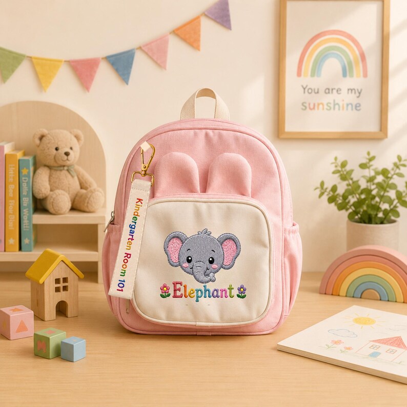 K&ouml;nnte beinhalten: Ein hellrosa Rucksack mit zwei abgerundeten Ohren an der Oberseite und einer Vordertasche mit einem gestickten grauen Elefanten, dem Wort Elephant und zwei kleinen Blumen. Ein wei&szlig;er Stoffanh&auml;nger am Rei&szlig;verschluss tr&auml;gt die Aufschrift Kindergarten Room 101.