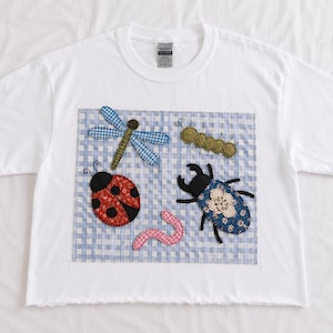 May include: A white short-sleeve t-shirt featuring a square patch with a light blue and white gingham background. The patch displays fabric appliqués of a blue dragonfly, a red ladybug, a green caterpillar, a pink worm, and a blue beetle.